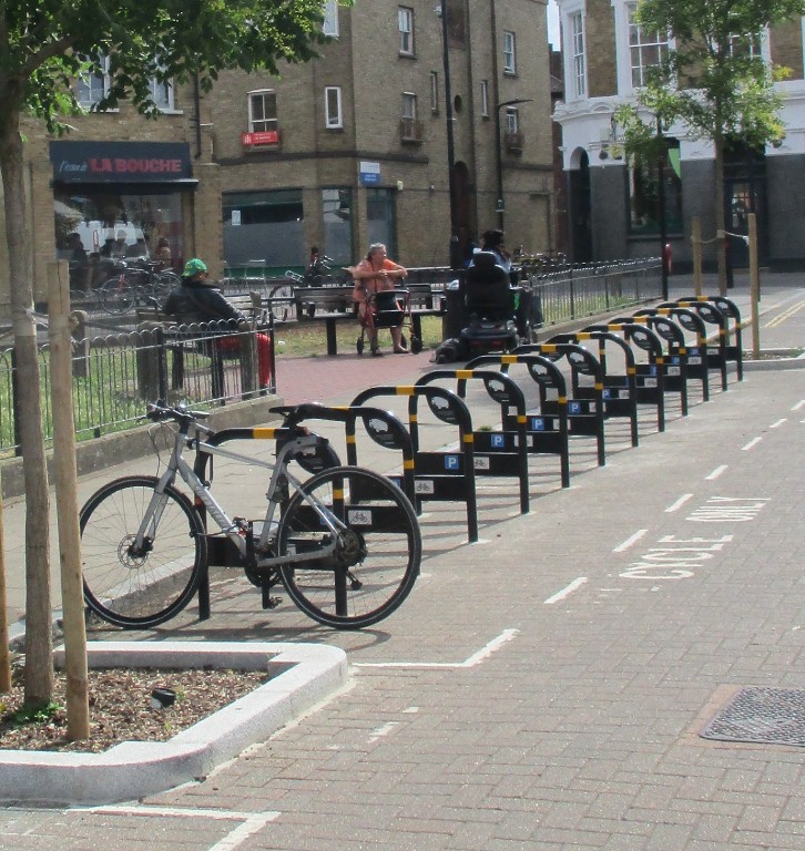Outdoor cycle stands on a carriageway with several empty spaces, a lone bicycle parked, and a person on a scooter passing by.